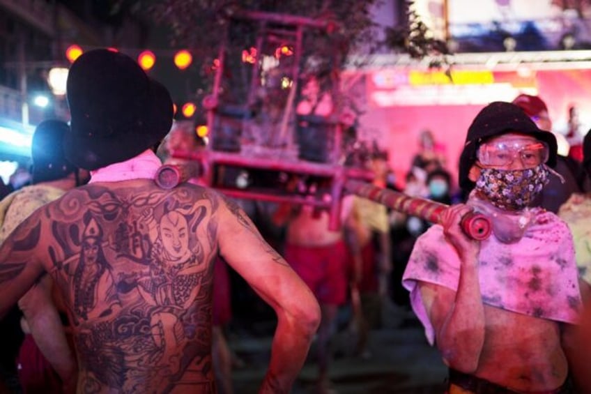 Four men donning only red shorts and protective eyegear carry the palanquin holding Master