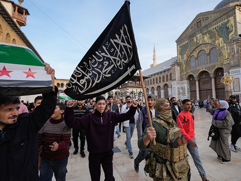 A masked opposition fighter carries a flag of Hayat Tahrir al-Sham (HTS) in the courtyard