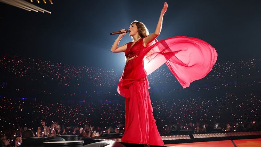 Taylor Swift puts her arm in the air wearing a red dress with part of it flying in the wind behind her