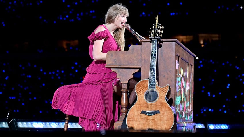 Taylor Swift in a pink dress plays behind a brown piano with a guitar leaning up against it at the Eras Tour
