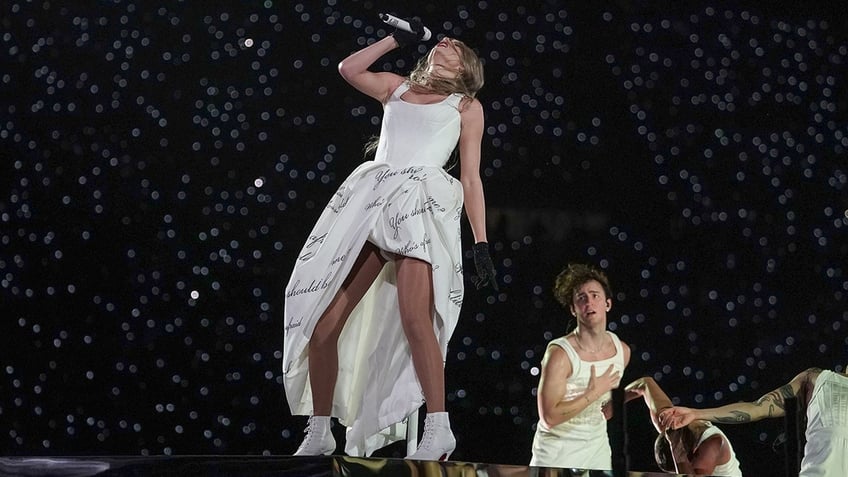 Taylor Swift in a white dress tilts her head back and sings into her microphone