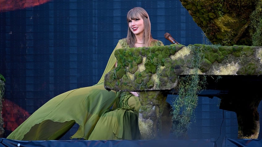Taylor Swift in a green dress sitting behind her mossy piano smiles at the crowd