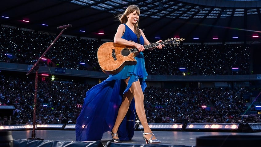 Taylor Swift in a long blue dress struts the stage while playing the guitar in Edinburgh 