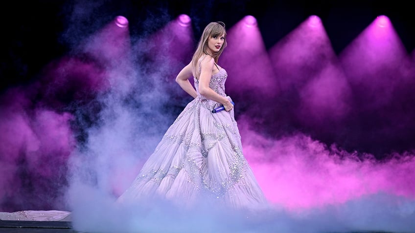 Taylor Swift in a purple dress looks back at her audience while on stage
