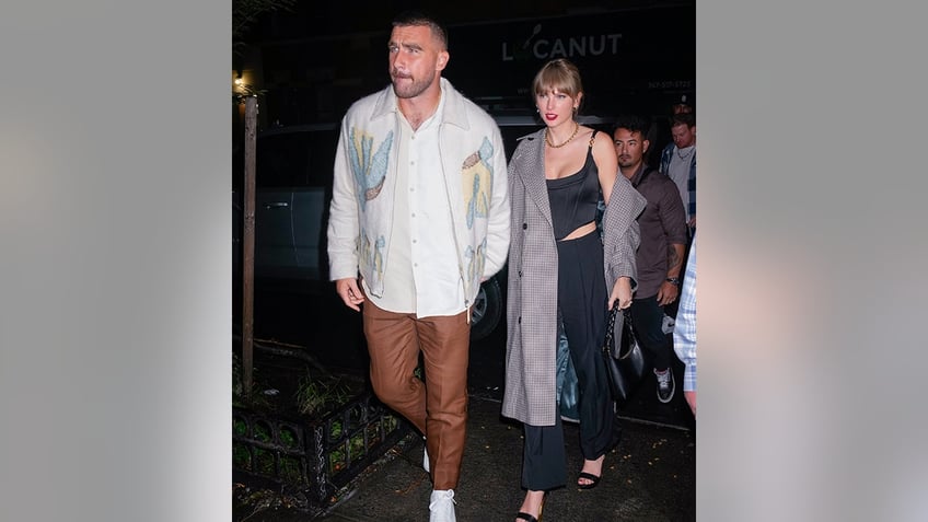 Travis Kelce in a white printed shirt and brown pants leads Taylor Swift in black and a grey coat in New York City