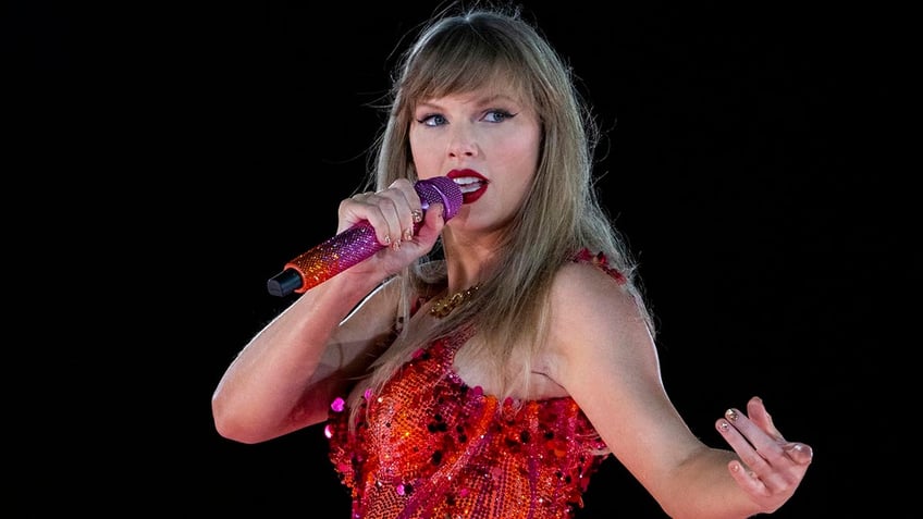 Taylor Swift sings into an orange and purple bedazzled microphone looking to her left on stage wearing a red/orange bodysuit