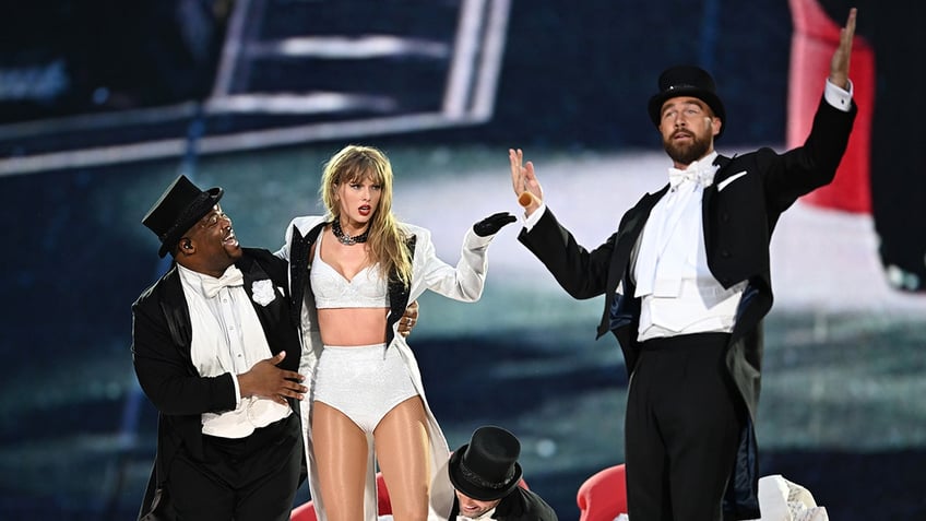 Taylor Swift in her white two piece outfit and white jacket performs on stage with backup dancer Cam and boyfriend Travis Kelce, donning a top hat
