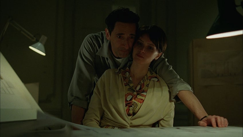 Adrien Brody and Felicity Jones standing close together in a scene from "The Brutalist"