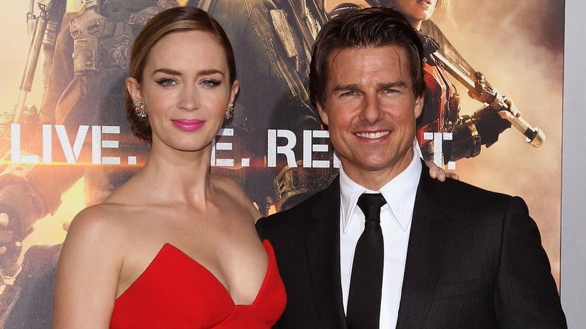 Emily Blunt in a red strapless dress soft smiles on the carpet with Tom Cruise in a black suit and tie