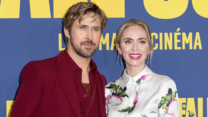 Ryan Gosling in a dark red shirt and suit looks stoic on the camera with a smiling Emily Blunt in a white patterned dress