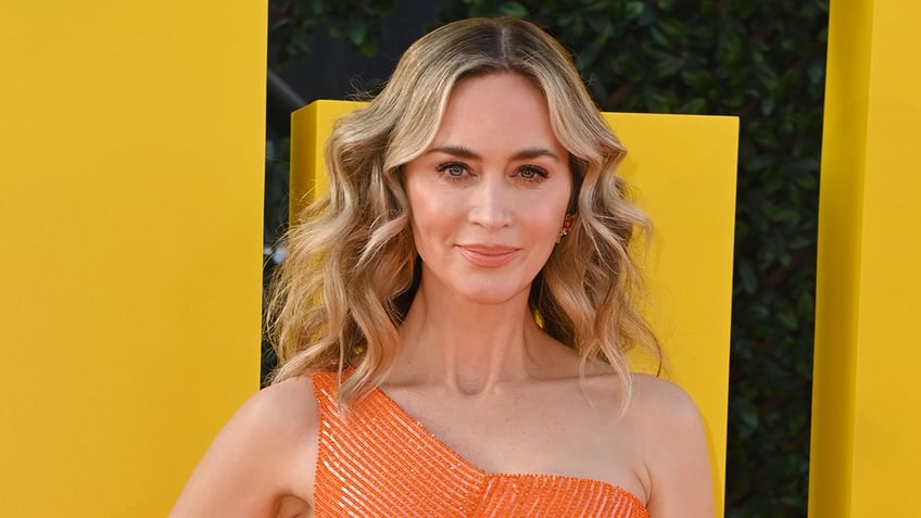 Emily Blunt soft smiles on the carpet in a sparkling orange one-shoulder dress