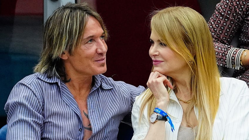 Keith Urban in a blue and white striped shirt looks up as Nicole Kidman with her hand under her chin looks adoringly at her husband at the US Open