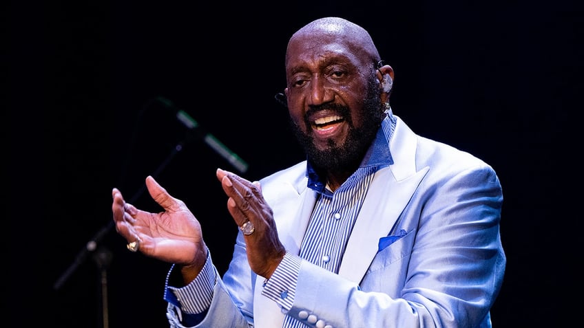 Otis Williams wearing a pale blue and white suit singing on stage.