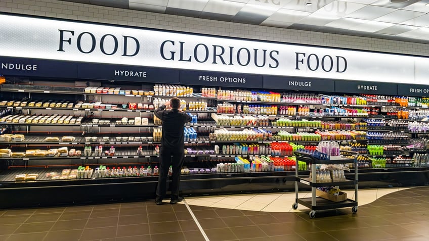 A man stocks drinks in the refrigerated section of a convenience store under a sign that reads "Food Glorious Food."