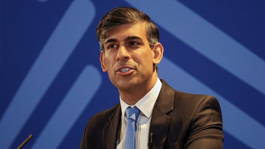 British Prime Minister Rishi Sunak stands in front of a microphone wearing a dark suit, white shirt, and blue tie in front of a blue backdrop.
