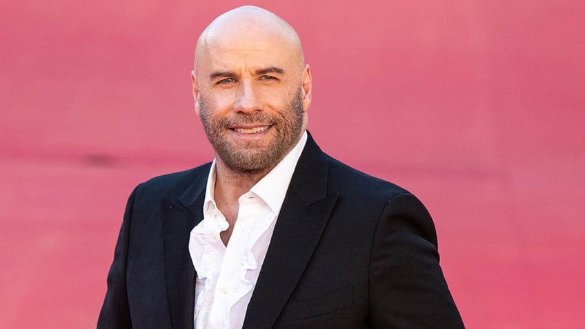 John Travolta in a black suit and white shirt smiles on the carpet