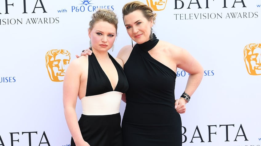 Kate Winslet in a black dress poses with her hand on her hip with daughter Mia Threapleton in a black dress with a cream band around the waist on the carpet