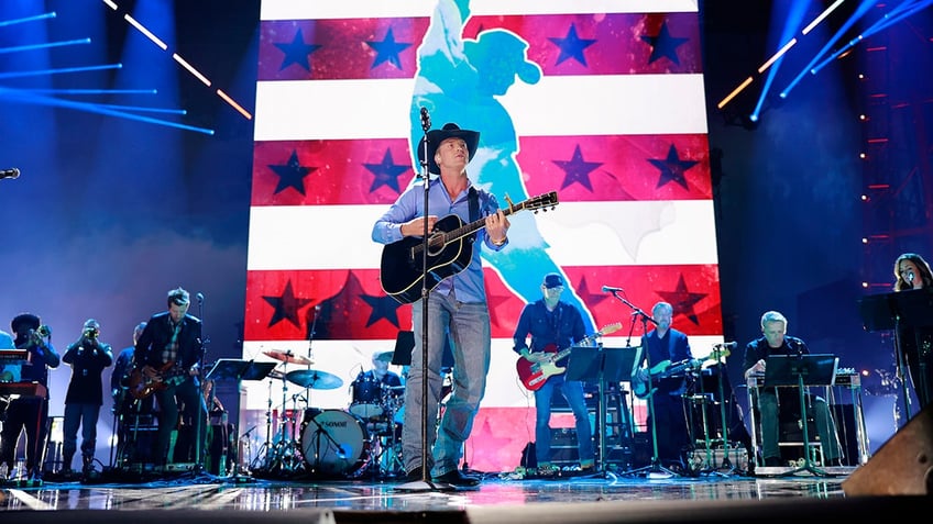 Parker McCollum in a blue shirt and jeans plays the guitar on stage for a Toby Keith tribute