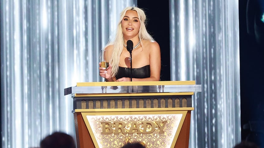 Kim Kardashian stands with champagne in her hand behind the podium at the Tom Brady roast