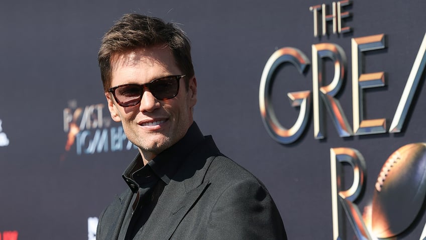 Tom Brady in a black suit and sunglasses looks dapper looking back at the camera at his roast 