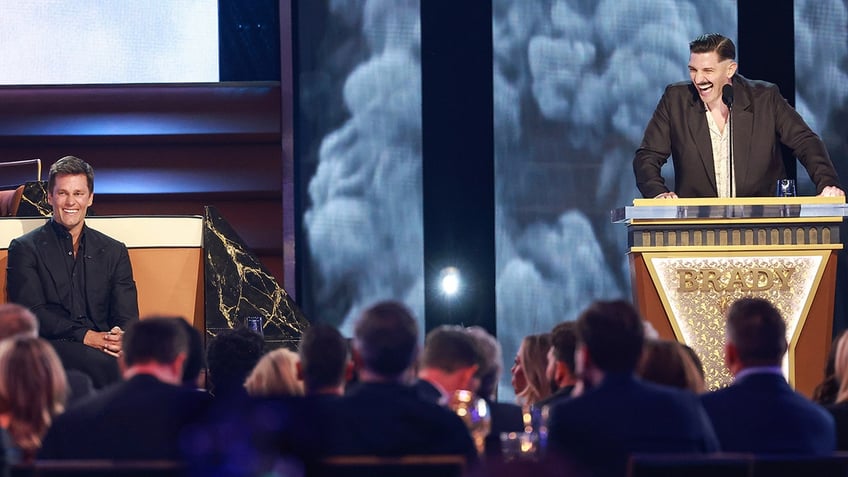 Tom Brady in a black suit sits on stage and smiles as he's being roasted by comedian Andrew Schulz behind a gold podium