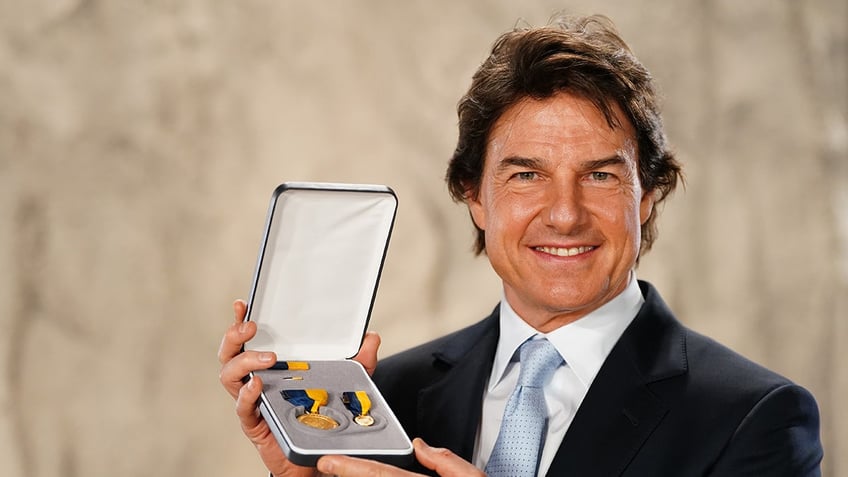 Tom Cruise wearing a black blazer a white shirt and a light blue tie holding a medal.