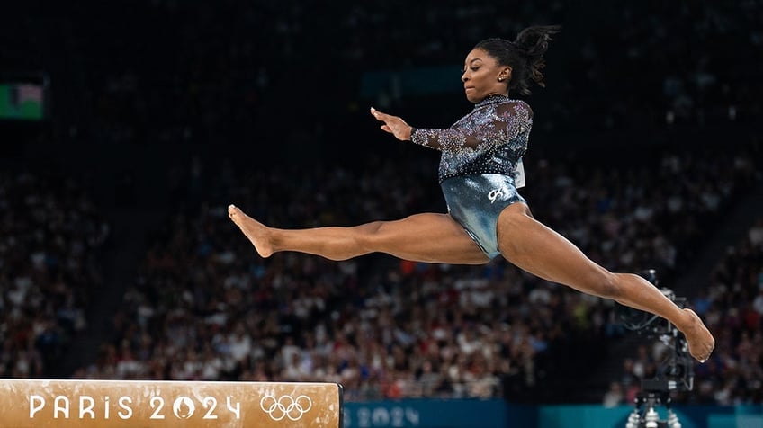 Simone Biles with legs spread apart leaping onto the balance beam