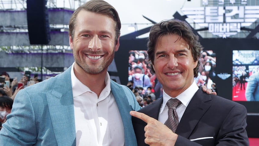 Glen Powell wearing a white button down and blue/teal blazer poses next to a pointing Tom Cruise in a black suit on the red carpet
