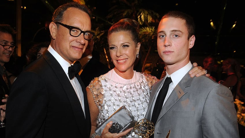 Tom Hanks in a tuxedo and black rimmed glasses smirks at the camera next to wife Rita Wilson in a white dress next to son Chet in a grey suit and black tie