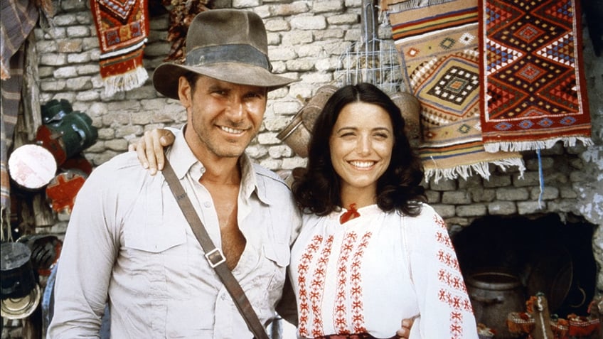 Harrison Ford in a white shirt smiles next to Karen Allen in a white blouse on the "Indiana Jones" set
