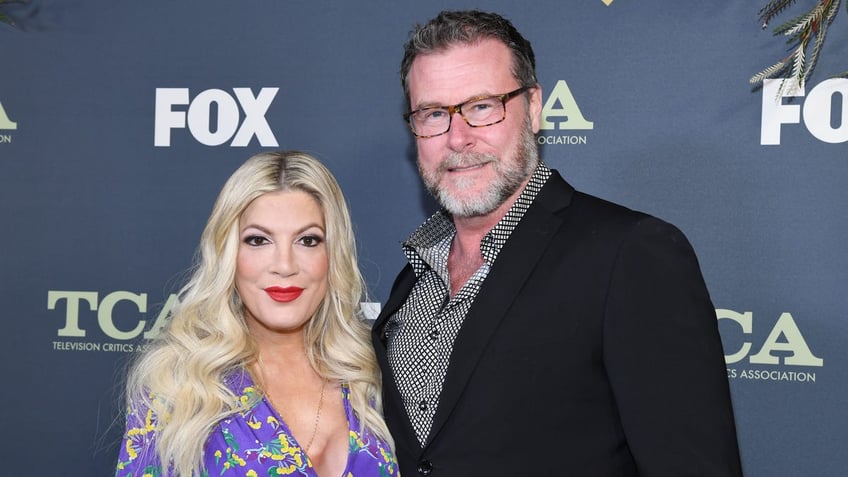 Tori Spelling in a purple blouse smiles with husband Dean McDermott in a black jacket on the carpet