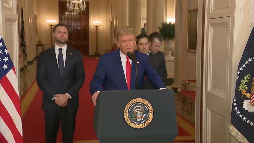 President Trump speaks at a lectern bearing the presidential seal, flanked by officials in the White House Cross Hall.