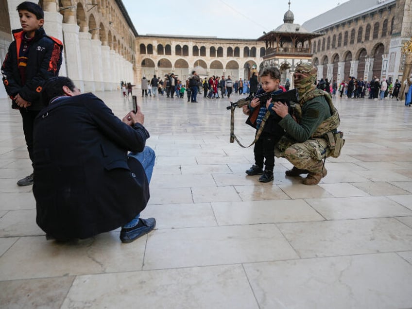 A boy poses for a photo with a masked Hayat Tahrir al-Sham (HTS) fighter holding a gun in