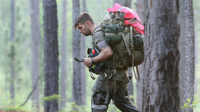 A soldier walks through the woods with a large backpack and checks a compass while completing a land navigation course during Special Forces Assessment and Selection
