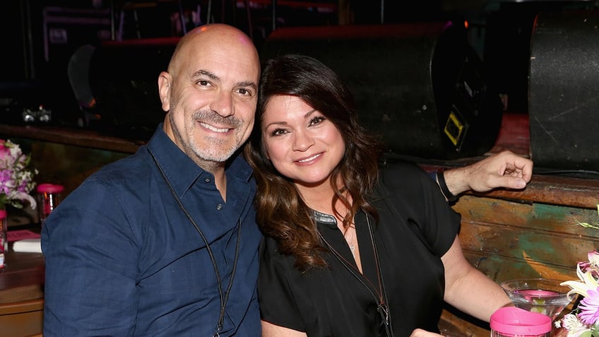 Tom Vitale smiles wearing a blue shirt as wife Valerie Bertinelli in black leans into him.