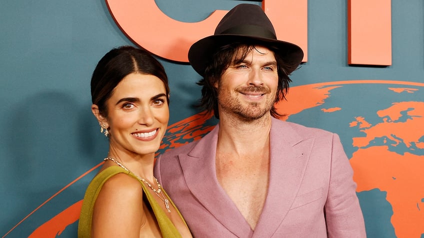 Nikki Reed in a green dress smiles at the camera with husband Ian Somerhalder in a light pink blazer with no shirt underneath and a brown hat