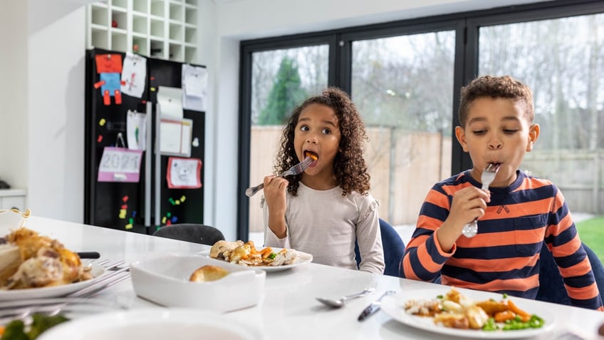 A girl and a boy sit at a kitchen counter eating meat with forks.