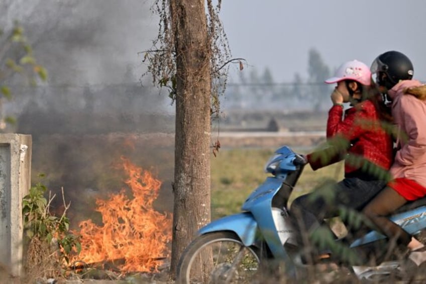 A woman covers her face as she rides a motorbike past a burning garbage dump on the side o