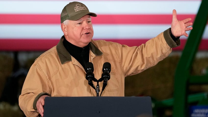 Gov. Walz in tan jacket and olive green hat speaking at lectern