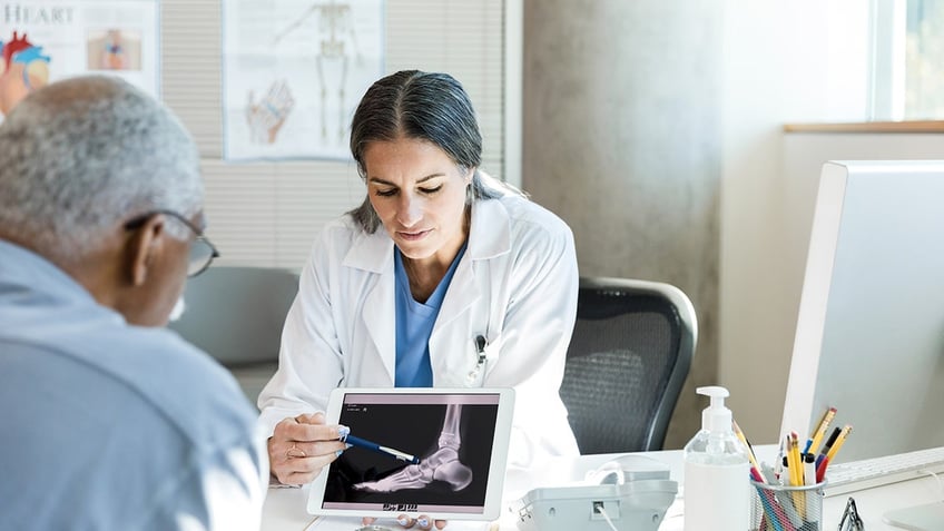 orthopedic doctor points to something on a senior male patient's foot x-ray.