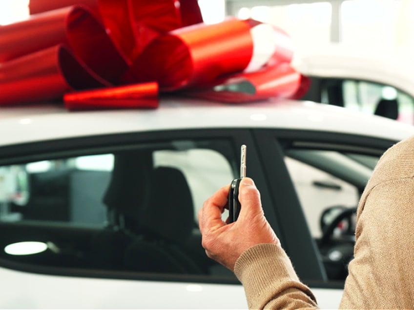 The man stands facing the automobile and the camera takes it off the back. He looks at the car and smiles. The car is tied with a big red bow. Then he takes out the key to this car