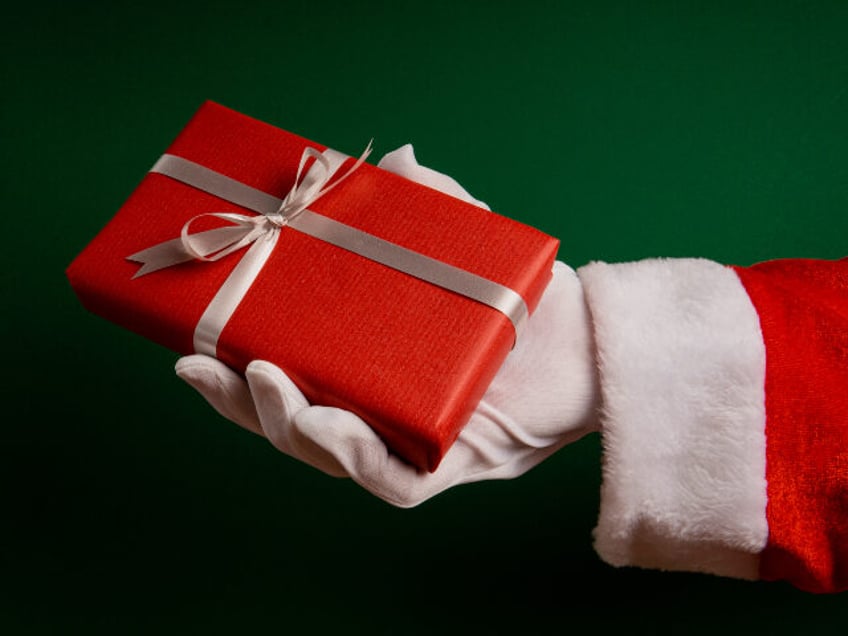 Hand of santa claus gives a red wrapped box with a silver ribbon on a green background wit