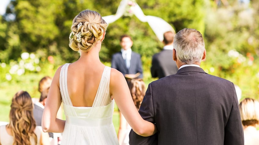 Rear view of bride and father walking down the aisle with people in background during outdoor wedding.