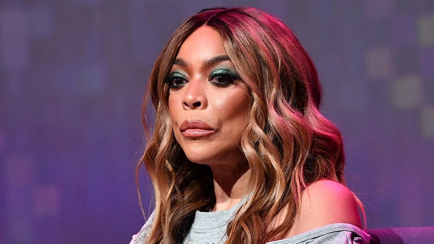 Wendy Williams in a grey shirt with a shoulder cut out looks down and downcast on stage