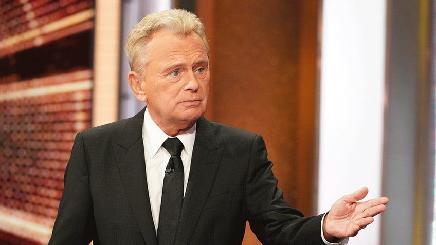 Pat Sajak in a black suit and tie looks off in the distance on an episode of "Wheel of Fortune"