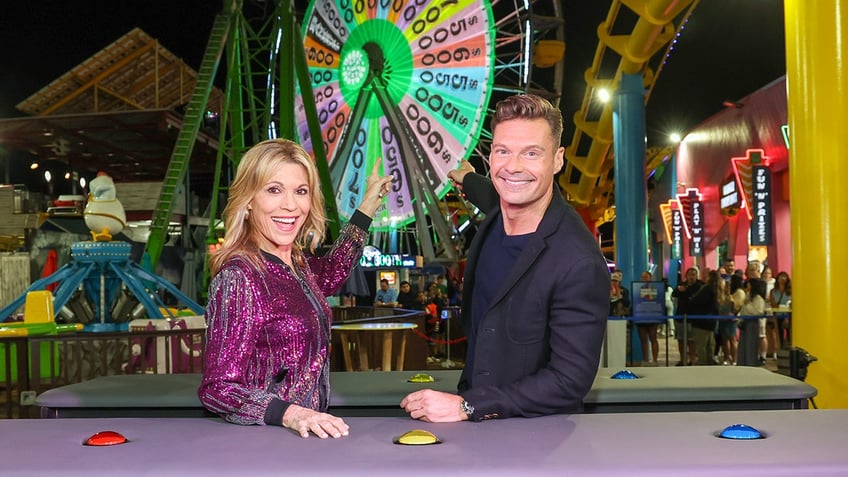 Vanna White and Ryan Seacrest point at the Santa Monica ferris wheel lit up like "Wheel of Fortune" wheel