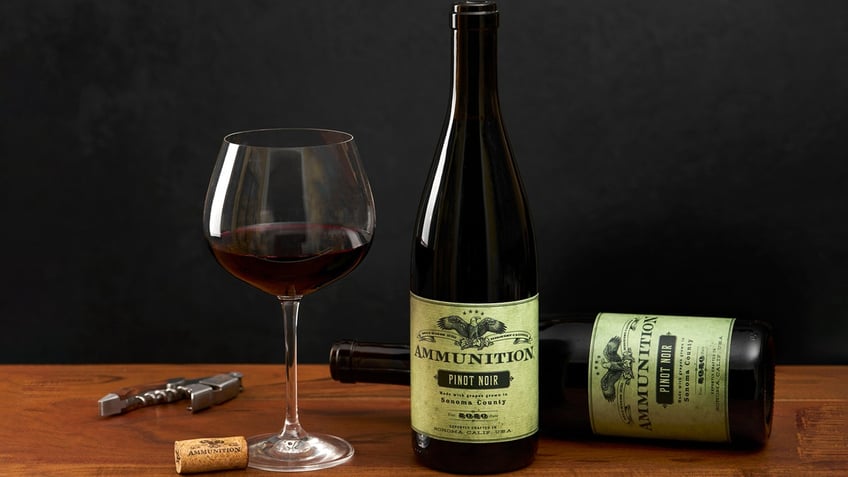 A glass of red wine is visible on a table next to two bottles of Ammunition's pinot noir.