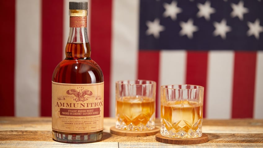 A bottle of Ammunition whiskey is shown on a wooden table next to two glasses with an American flag as a backdrop.