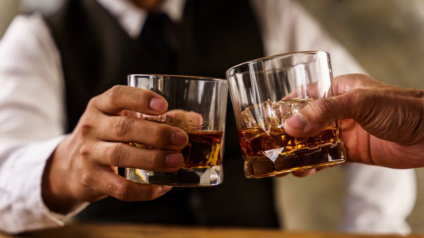 Two hands holding bourbon glasses touch as a gesture of cheers.