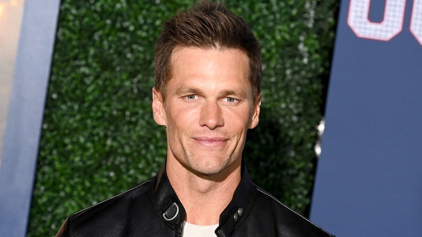 Tom Brady in a white shirt and leather jacket soft smiles and stares right at the camera on the "80 For Brady" red carpet